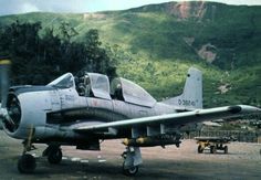 a fighter jet sitting on top of a tarmac next to a forest covered hillside