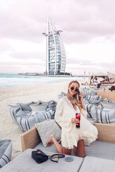 a woman sitting on top of a beach next to the ocean with a drink in her hand