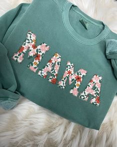 a green sweatshirt with flowers and the word mama on it is laying on a furry surface