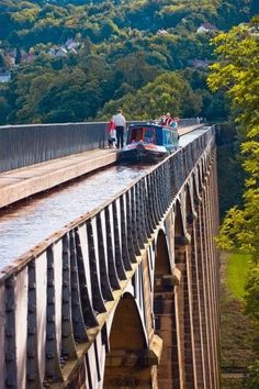 people are standing on the side of a bridge with a boat going over it and trees in the background