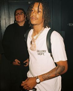 a man with dreadlocks wearing a white shirt and carrying a black bag while standing next to another man