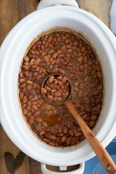 a wooden spoon in a white pot filled with baked beans and brown liquid on top of a wood table