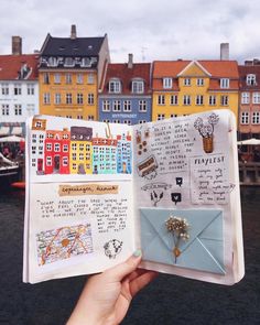 someone is holding up an open book with pictures and writing on it in front of some buildings