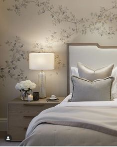 a bed with white sheets and pillows next to a night stand, lamp and flowers