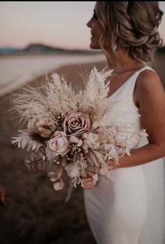 a woman in a white dress holding a bouquet of flowers and feathers on the beach