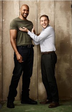 two men standing next to each other in front of a wooden wall with one holding the other's hand