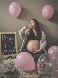 a pregnant woman sitting on the floor drinking from a bottle while surrounded by balloons and signs