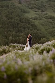 a bride and groom standing on top of a lush green hillside surrounded by wildflowers