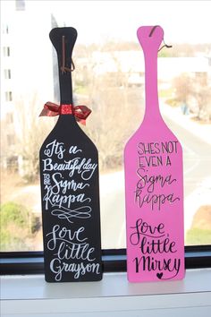 two wine bottles are sitting on a window sill, one has a ribbon and the other is painted pink