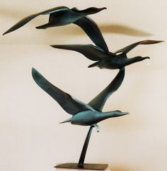 three metal birds flying in the air on top of a wooden base with a white wall behind them