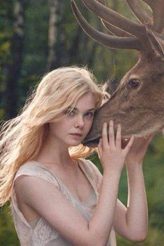 a beautiful blonde woman kissing a deer in the middle of an outdoor forest area with trees and bushes behind her