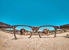 two people wearing hats and sunglasses laying on the sand with palm trees in the background