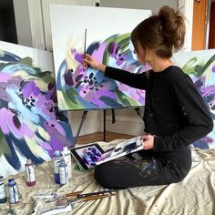 a woman sitting on the floor in front of two paintings and holding a paintbrush