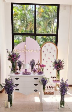 a white table topped with lots of purple flowers next to a window filled with plants