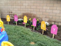 some pink and yellow signs sitting in the grass near a brick wall with flowers on it