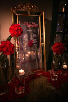 red roses in vases and candles on a table with a framed poster behind them