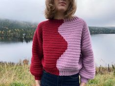 a woman standing in front of a lake wearing a pink and red sweater