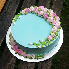 a blue cake with pink and white flowers on it sitting on a wooden table outside