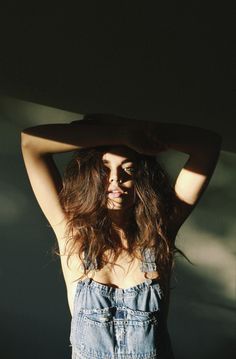 a woman with long brown hair and blue overalls is posing for the camera while holding her hands behind her head