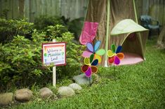 a sign that is in the grass near some rocks and flowers with a tent behind it