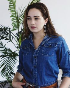 a woman standing in front of a potted plant wearing a denim shirt and brown belt