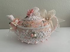 a pink lace covered tea pot with flowers on the top and bows in the bottom