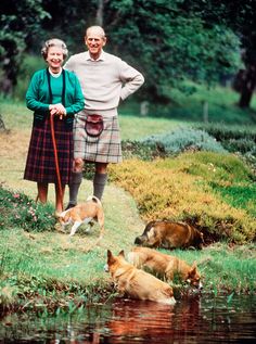 an older man and woman standing next to two dogs in the grass near some water