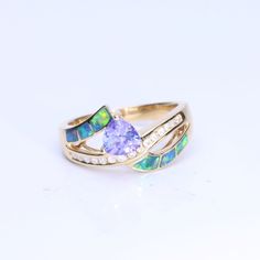 Wear the CIRARI 14K Yellow Gold Inlay Opal Trillion Tanzanite Ring to exude elegance. This ring is incredibly well-made and has a striking trillion-cut tanzanite set in a gorgeous inlay of opals. The design, set in dazzling 14K yellow gold, skillfully combines classic charm with contemporary refinement. This CIRARI masterpiece, a monument to timeless beauty and tasteful design, will elevate your look. Give yourself over to the allure of this magnificent ring. Trillion Cut Tanzanite Rings For Wedding, Fine Jewelry Tanzanite Rings With Diamond Cut, Elegant Tanzanite Trillion Cut Rings, Formal Trillion Cut Multi-stone Jewelry, Fine Jewelry Opal Ring With Multi-stone For Anniversary, Gold Trillion Cut Gemstone Ring, 14k Gold Jewelry With Trillion Cut Accent Stones, Tanzanite Trillion Cut Ring For Anniversary, Trillion Cut Tanzanite Ring For Anniversary