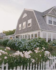 there is a white picket fence in front of a house with flowers on the side