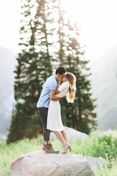 a man and woman kissing on top of a rock in front of pine trees with mountains behind them