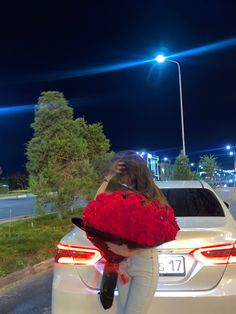 a woman holding a bouquet of red roses standing next to a white car at night