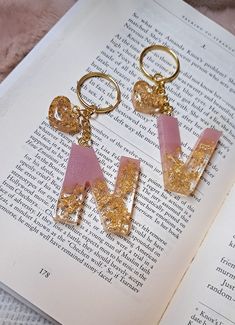 two pink and gold letters are on top of an open book, next to a pair of earrings