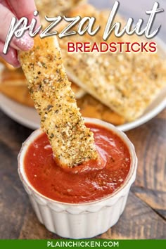 a hand dipping some bread sticks into a bowl of marinara sauce on a wooden table