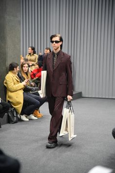 a man in a suit and sunglasses walking down the runway with shopping bags on his feet