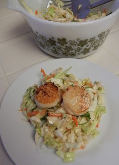 a white plate topped with scallops next to a bowl filled with coleslaw