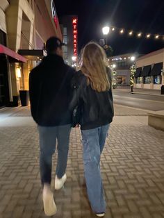 a man and woman walking down the street at night with their back to each other