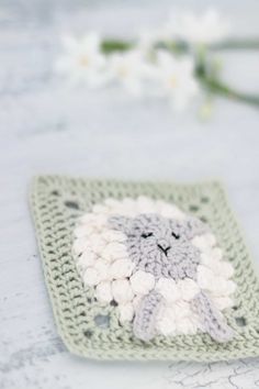 a crocheted square with a white and gray sheep on it next to flowers