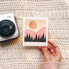 a person holding up a card with trees on it next to a polaroid camera