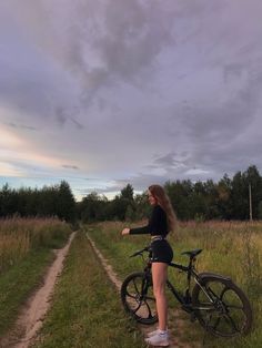 a woman standing next to a bike on a dirt road in the middle of a field