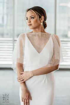 a woman wearing a white dress with sheer sleeves and an open back, standing in front of large windows