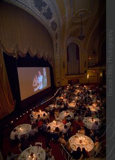 a large room filled with people sitting at tables and watching a movie on the screen