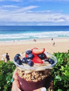 a person holding up a dessert in front of the ocean with strawberries and blueberries on top