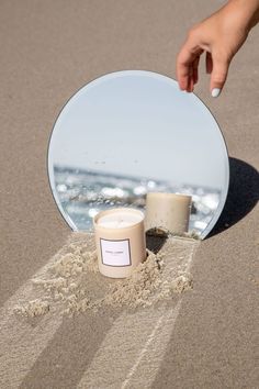 a candle sitting on top of a sandy beach next to a round mirror with a hand reaching for it