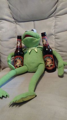 a stuffed frog sitting on top of a couch next to two beer bottles and a can