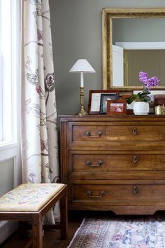 a bedroom with a dresser, mirror and lamp on it's side table in front of a window