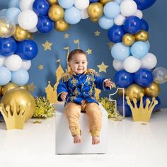 a baby sitting on top of a white box in front of blue and gold balloons