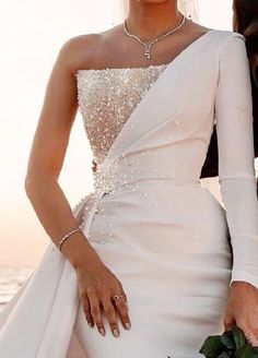 two women in wedding dresses standing next to each other near the ocean with their hands together