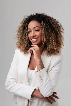 a woman with curly hair wearing a white suit and smiling at the camera while standing in front of a gray background