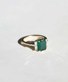 an emerald and diamond ring sitting on top of a white surface with one stone in the middle