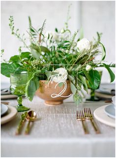 the table is set with white flowers and greenery in a copper vase on it